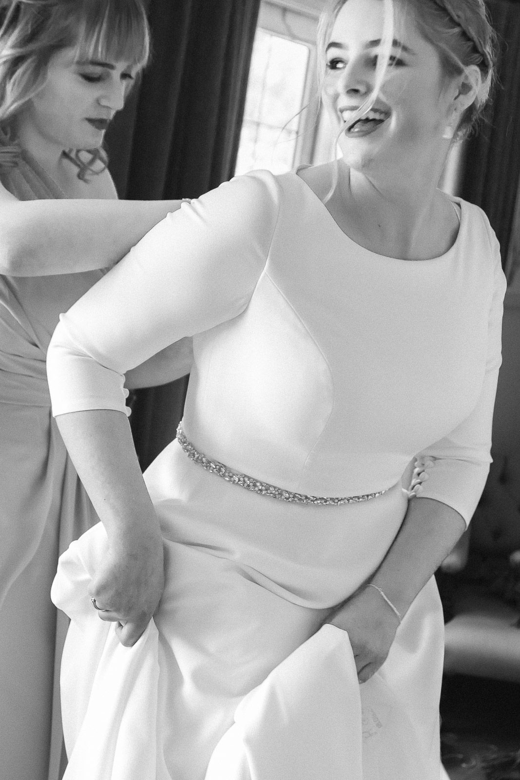Bride in elegant white gown with crystal belt being helped by bridesmaid, black and white wedding photo.