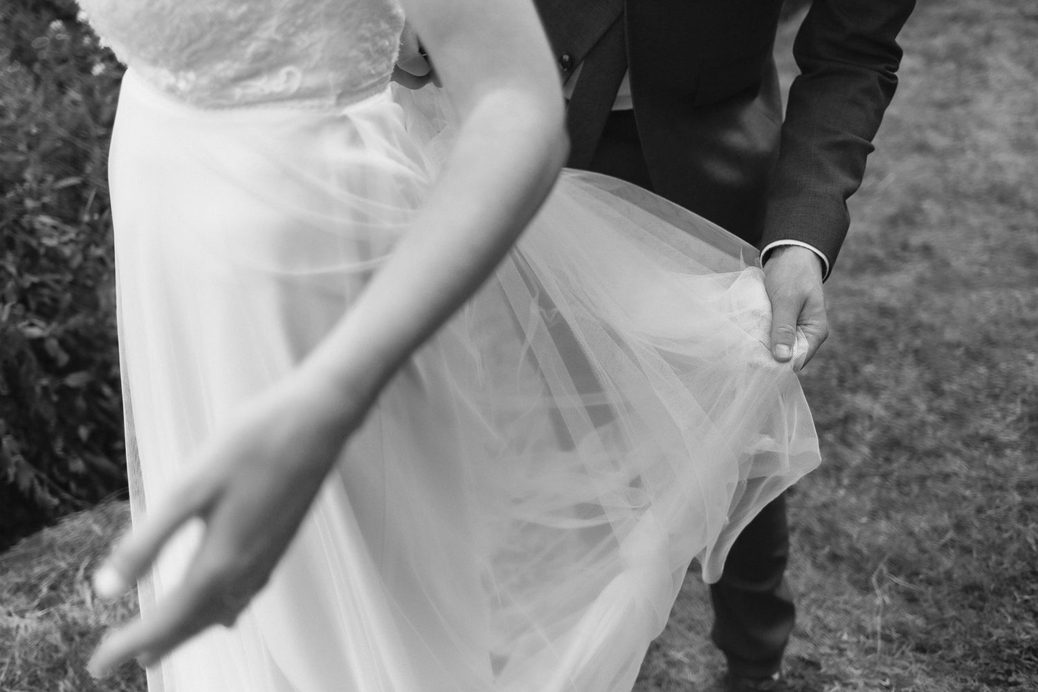 Black and white photo of bride and groom holding wedding dress tulle skirt outdoors.
