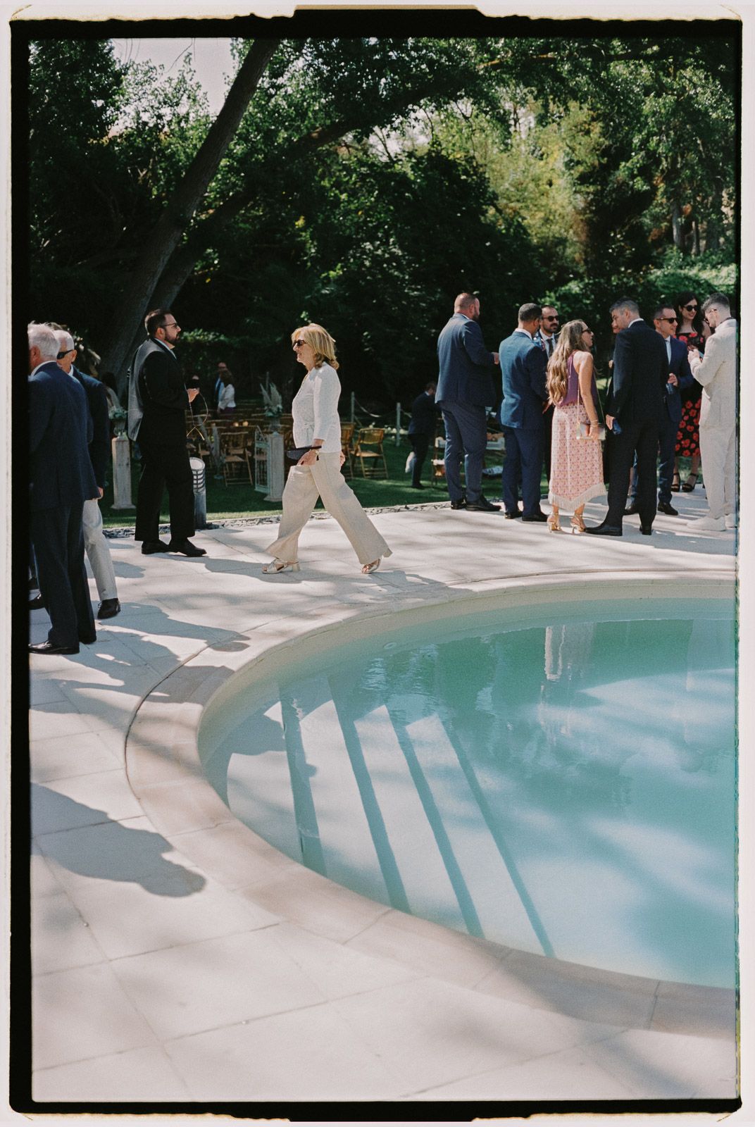 Guests mingle beside a round pool at an outdoor garden wedding reception on a sunny day.