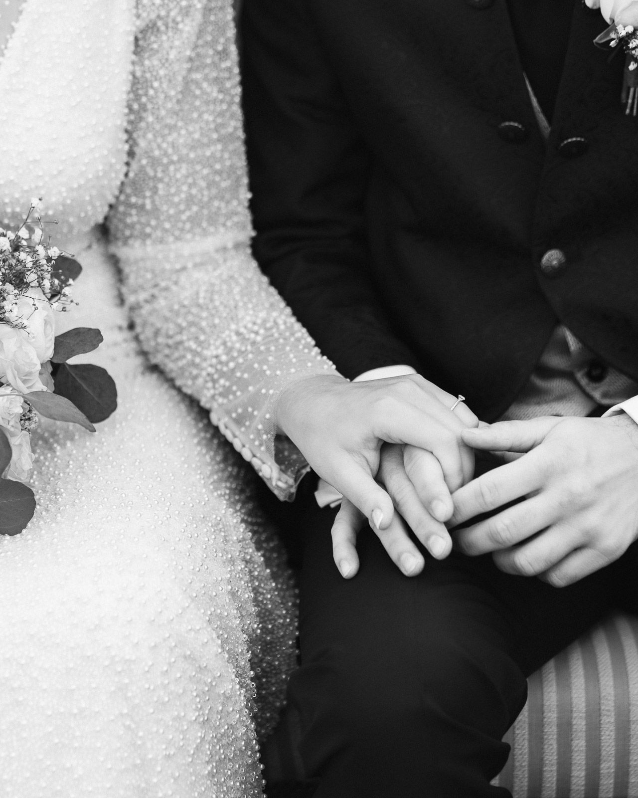 Black and white photo of a bride and groom holding hands, showcasing wedding rings and elegant attire.