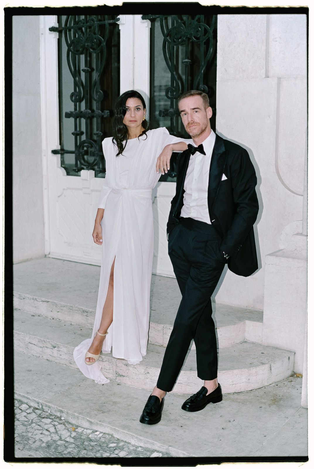 Elegant couple in wedding attire posing on steps, woman in white gown, man in black tuxedo.