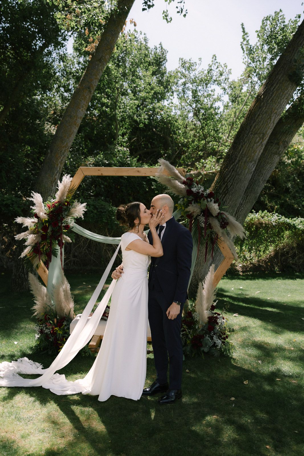 Bride and groom share first kiss under rustic hexagonal arch adorned with pampas grass at outdoor wedding.