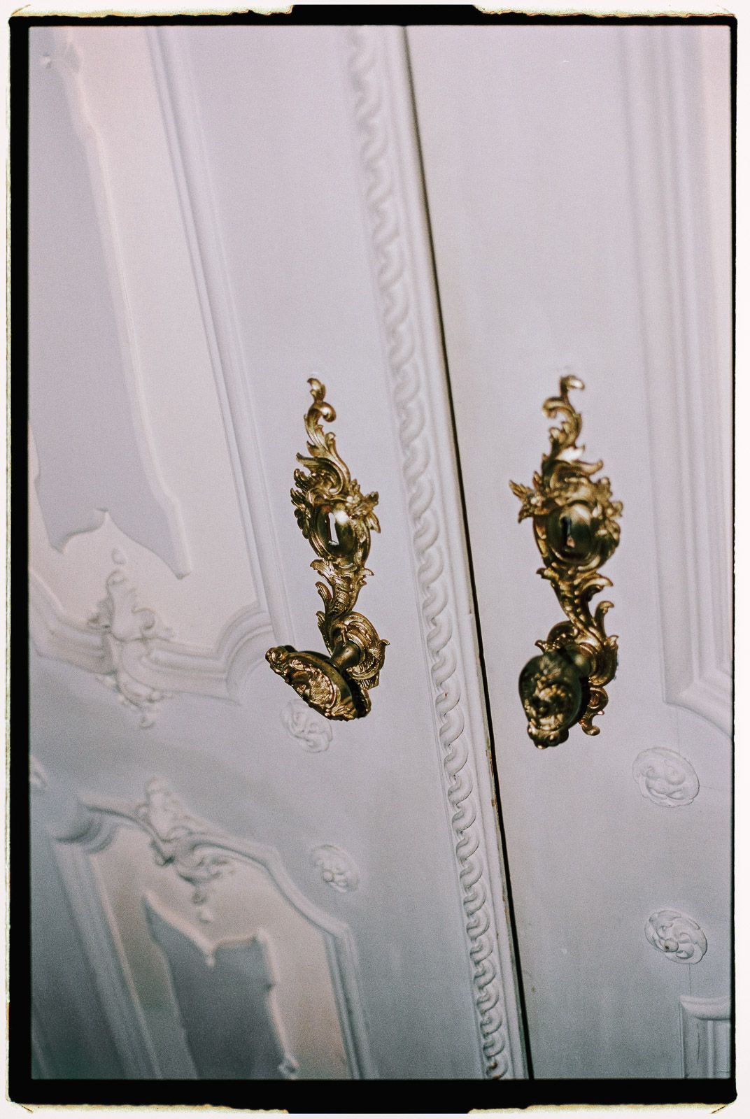 Ornate gold baroque door handles on white painted double doors with decorative molding.