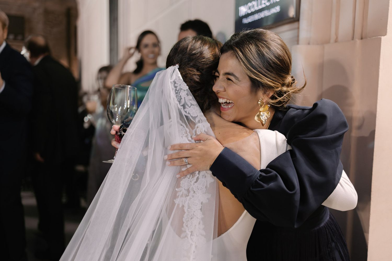 Bride in white gown and veil shares warm hug with guest in navy at elegant wedding reception.