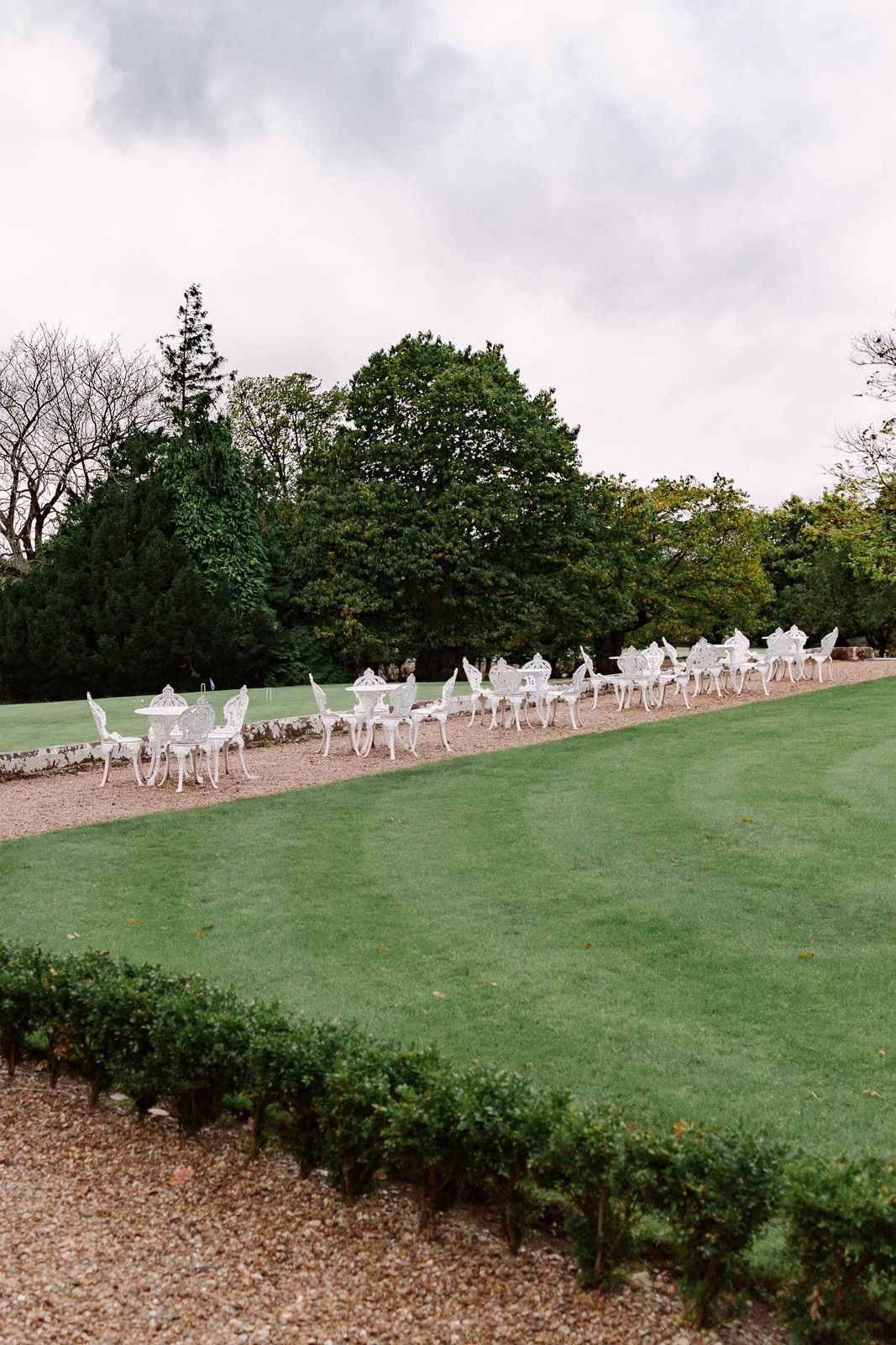 White garden chairs and tables arranged in rows on manicured lawn with trees backdrop.
