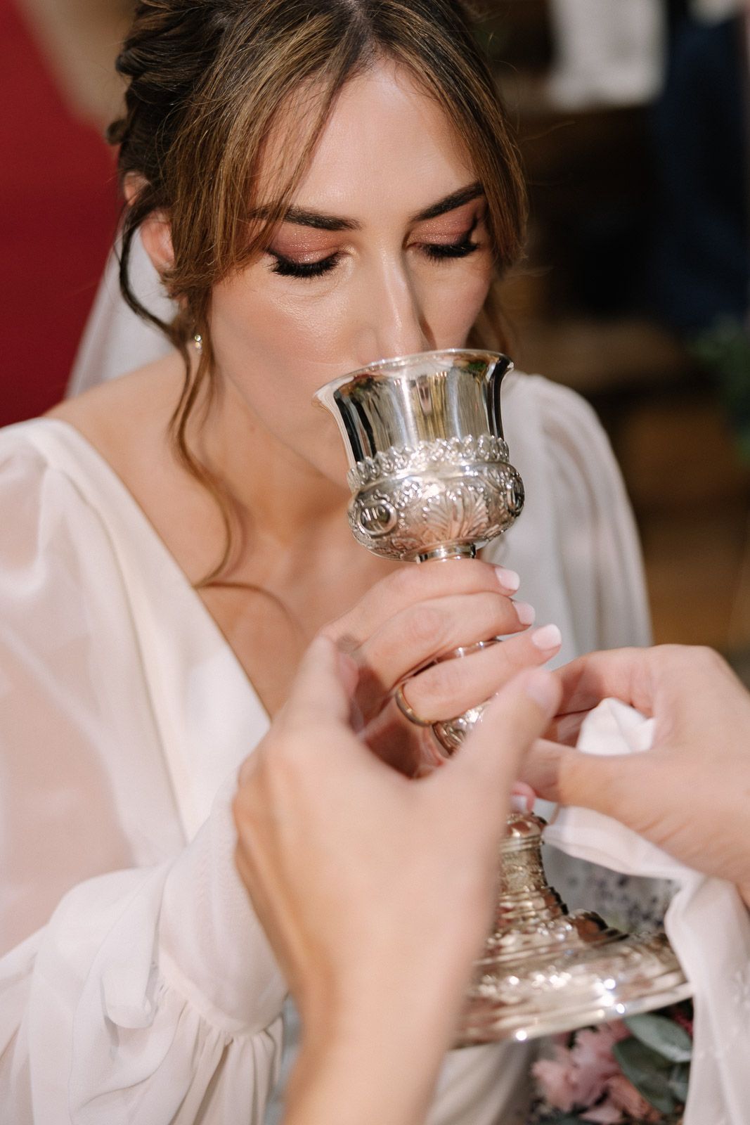 Bride in white dress drinking from ornate silver chalice during religious wedding ceremony.