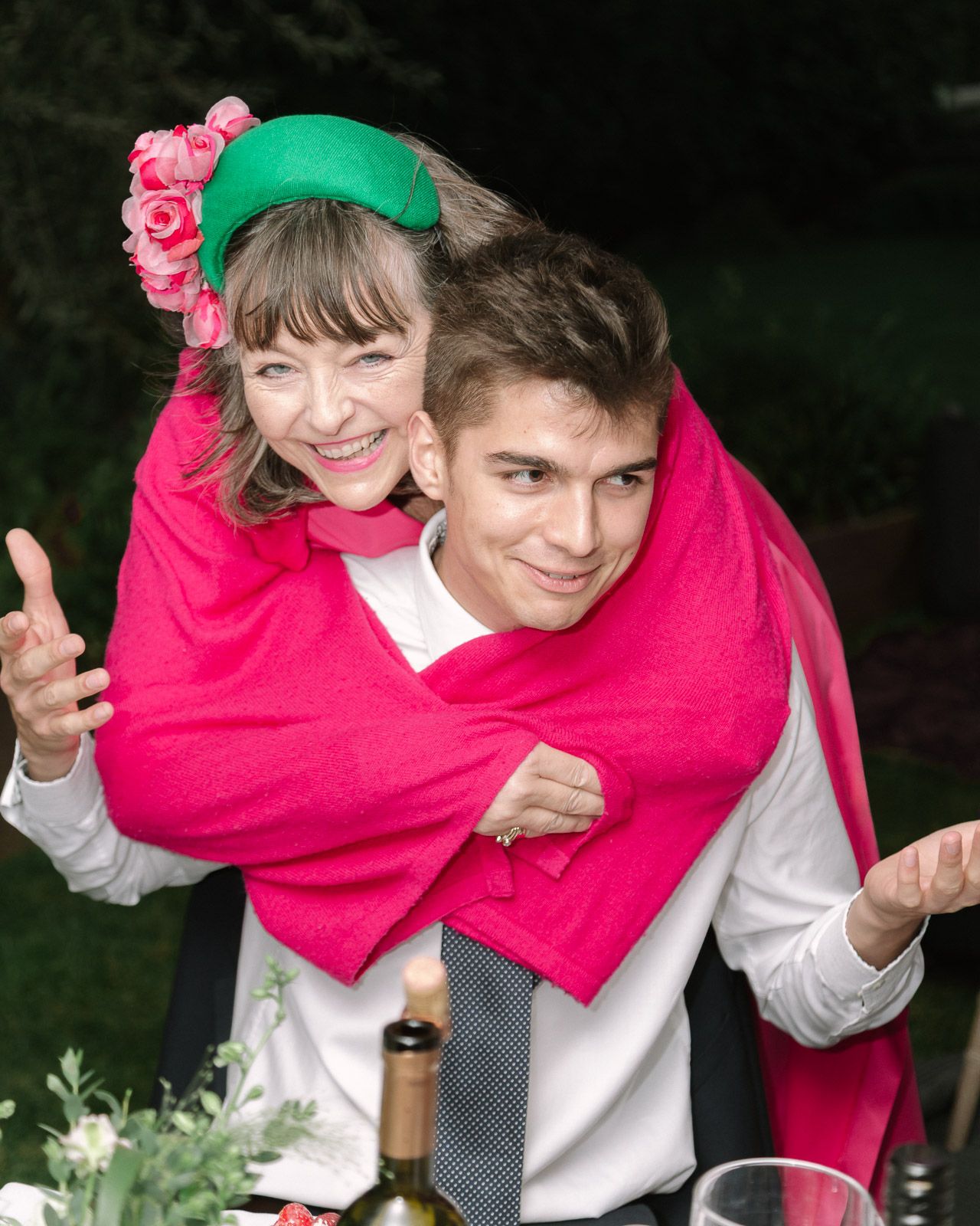 Woman in green floral hat and pink shawl hugging a young man in white shirt and tie at outdoor event.