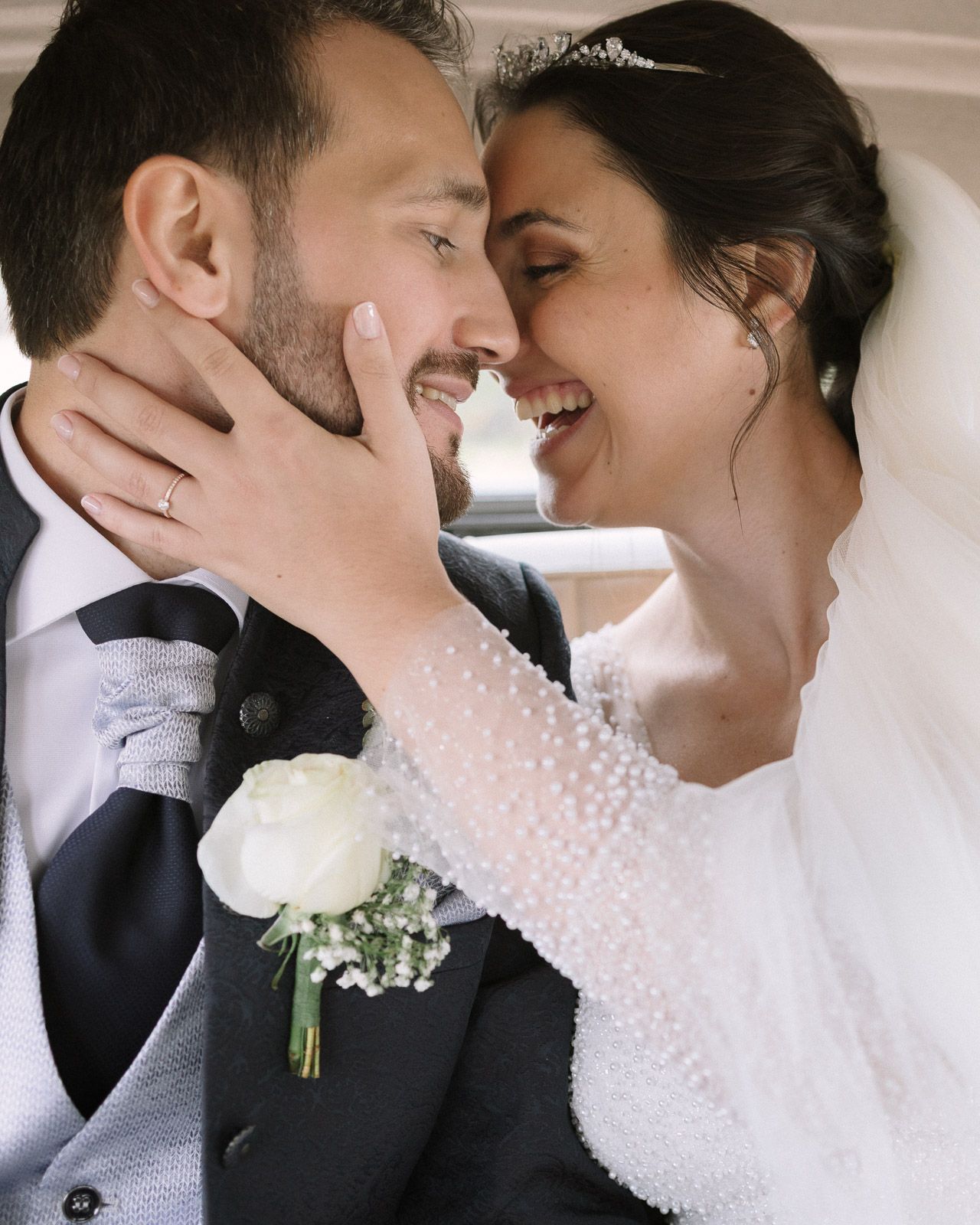 A bride and groom share an intimate moment, smiling at each other, with the bride in a pearl-adorned dress and veil.