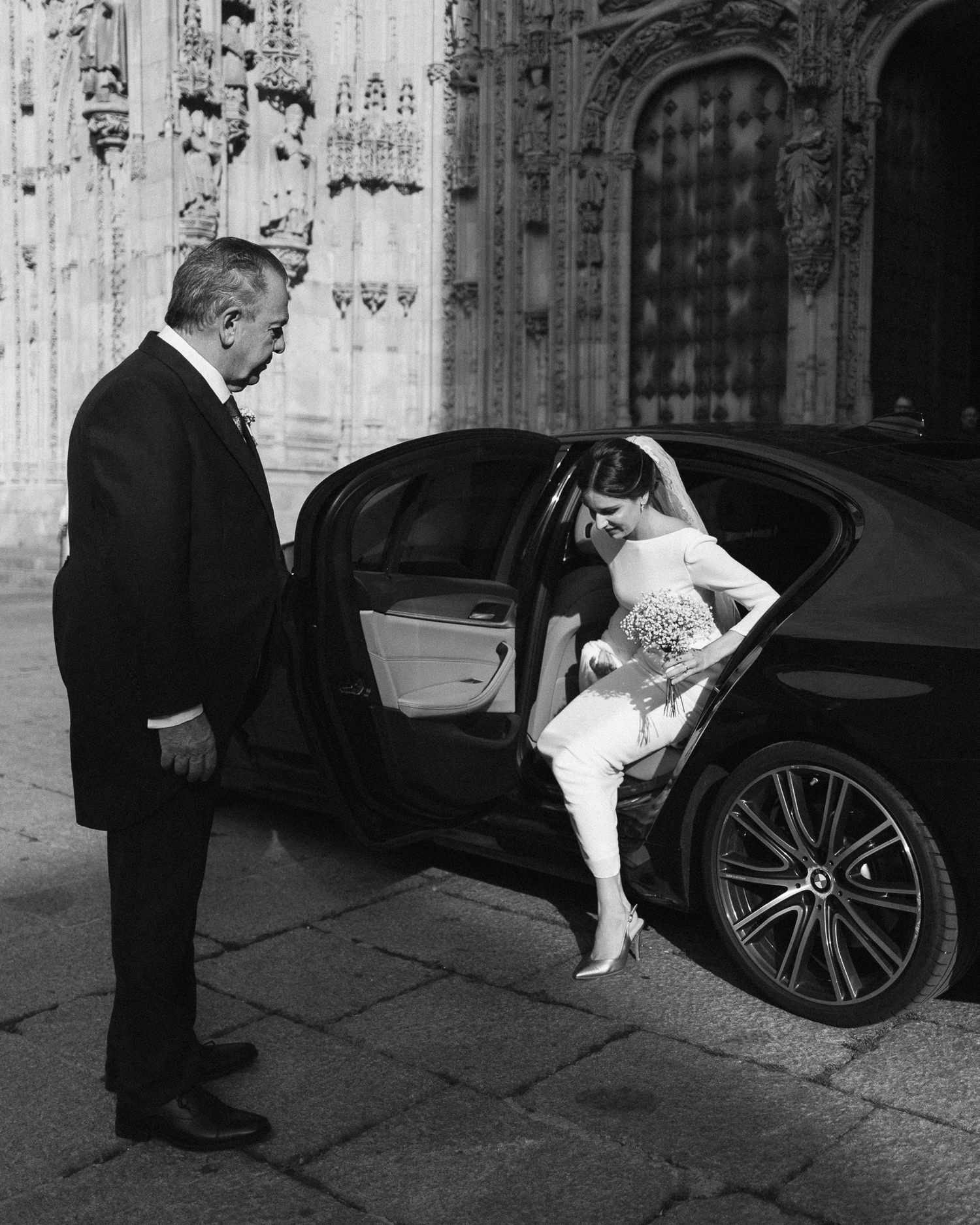 Formal black and white scene of a bride exiting a luxury car outside an ornate Gothic cathedral.