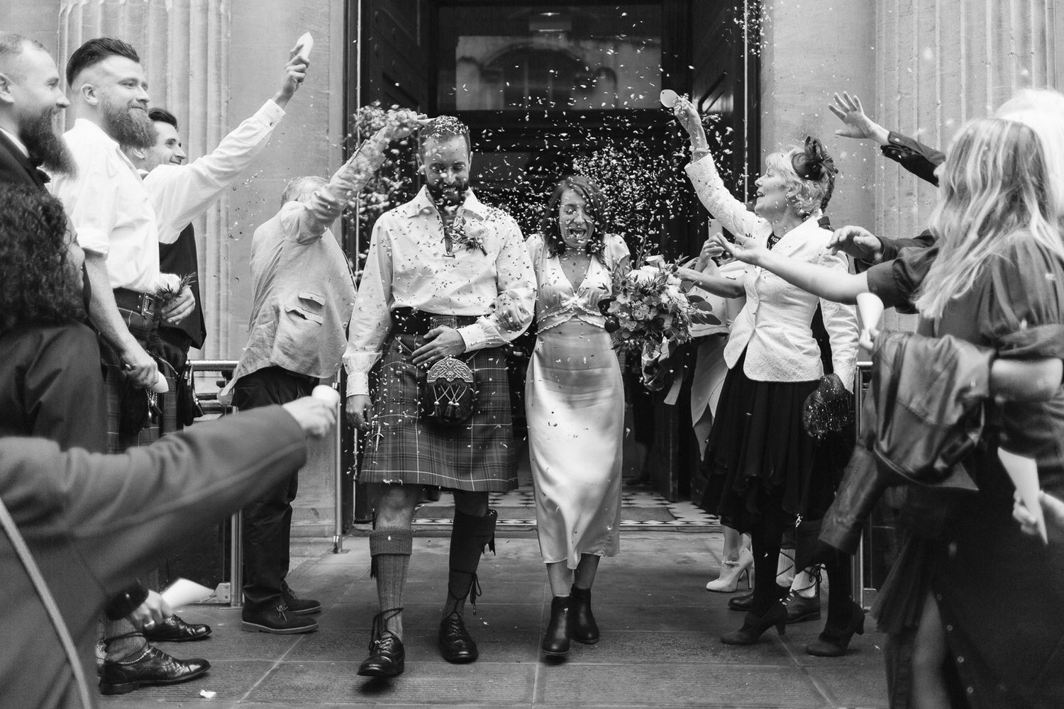 Black and white photo of a joyful couple in wedding attire exiting a church as guests throw confetti.
