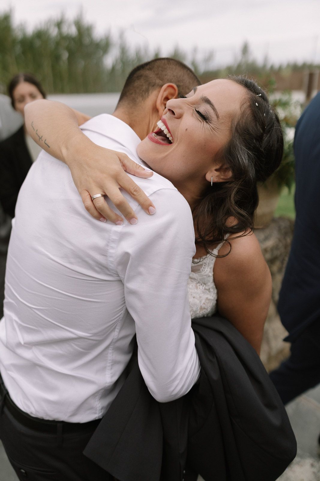 A joyful bride in a lace gown laughs while embracing a guest in a white shirt at an outdoor wedding celebration.