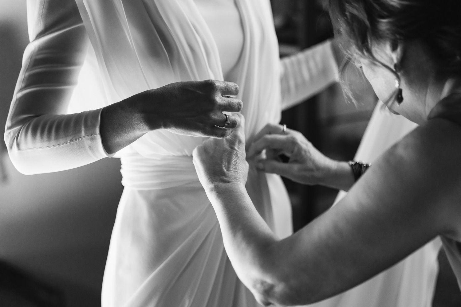 Black and white photo of bride getting wedding dress fastened by assistant before ceremony.