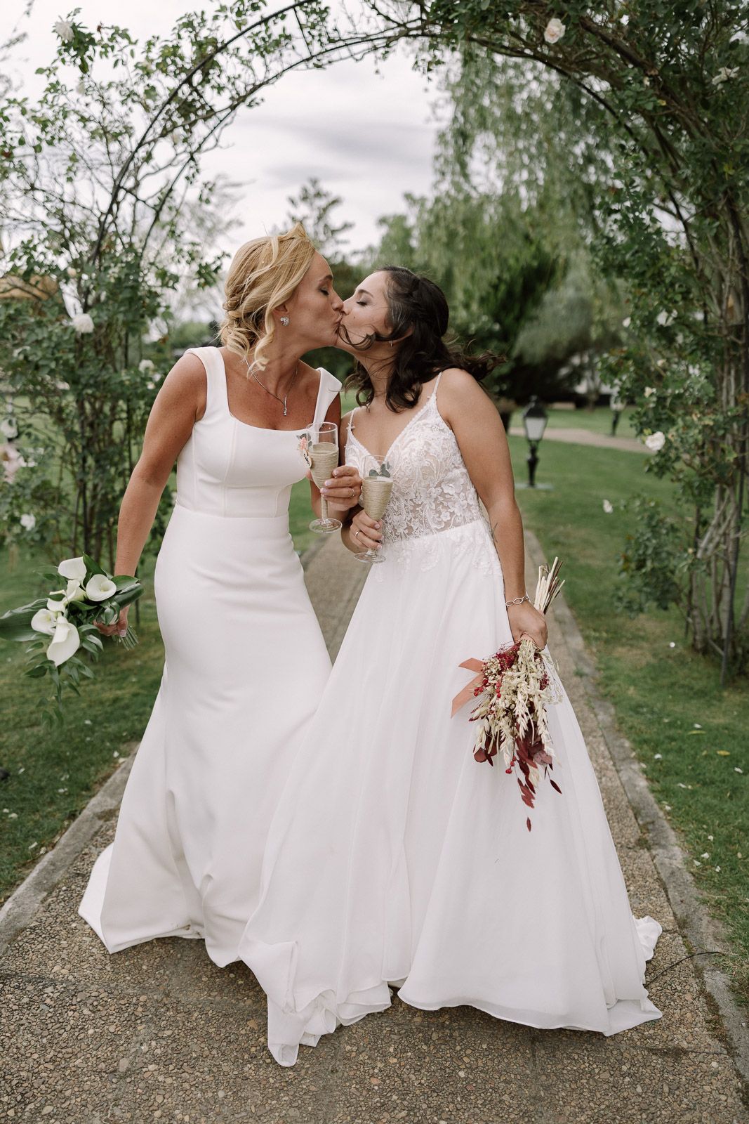 Two brides in white wedding gowns kissing under a floral arch while holding bouquets outdoors.
