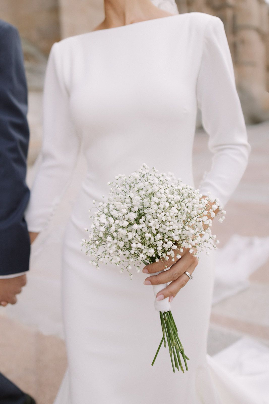 Bride in white long-sleeve wedding gown holding a delicate gypsophila bouquet with green stems at an outdoor ceremony.