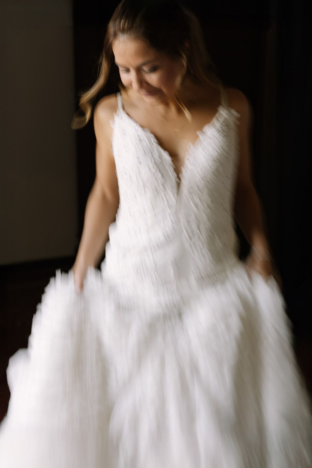Bride in white feathered wedding gown twirling in soft motion blur against dark background.