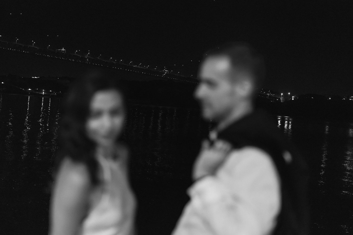 Blurry black and white photo of a couple near a bridge at night by the water.