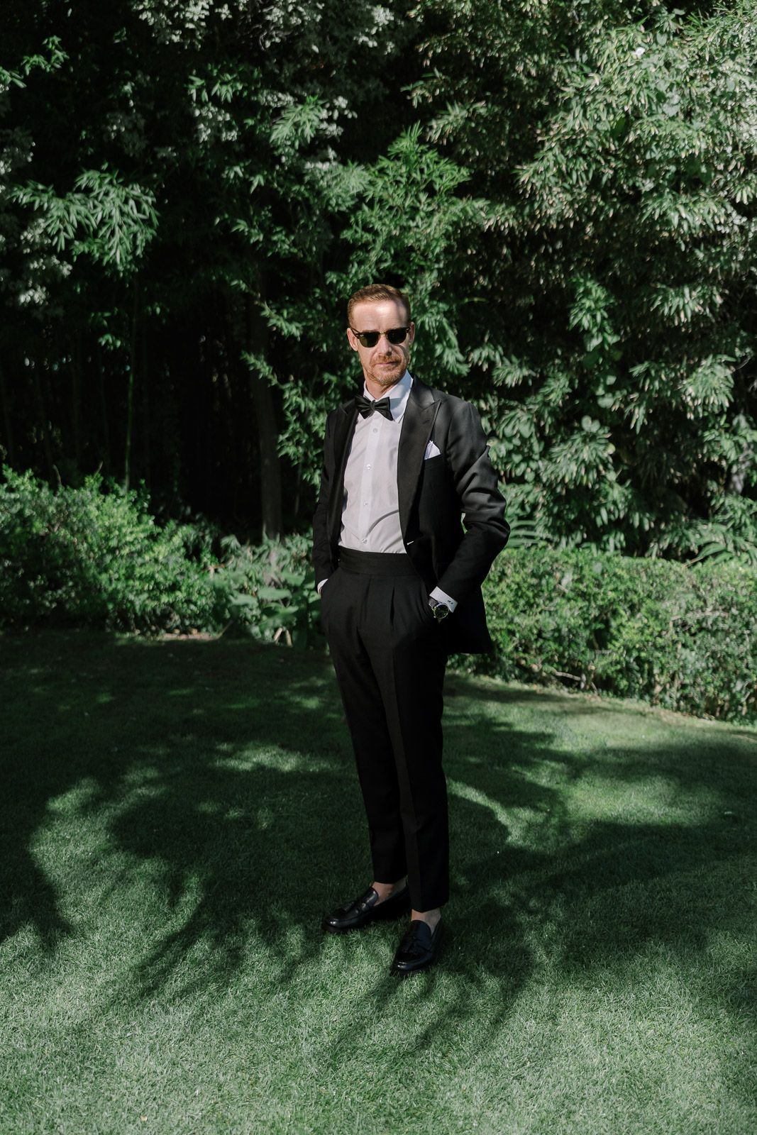 A dapper groom in a black tuxedo and sunglasses stands confidently on green grass with lush trees behind him.