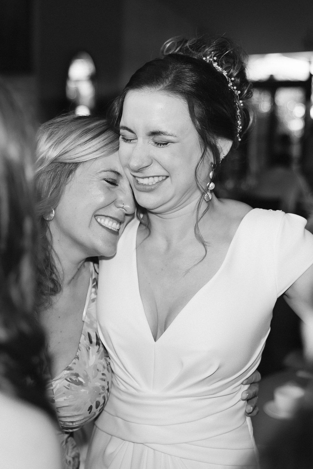 Two women laughing and embracing at a wedding, bride in white gown wearing a floral headpiece, black and white photo.
