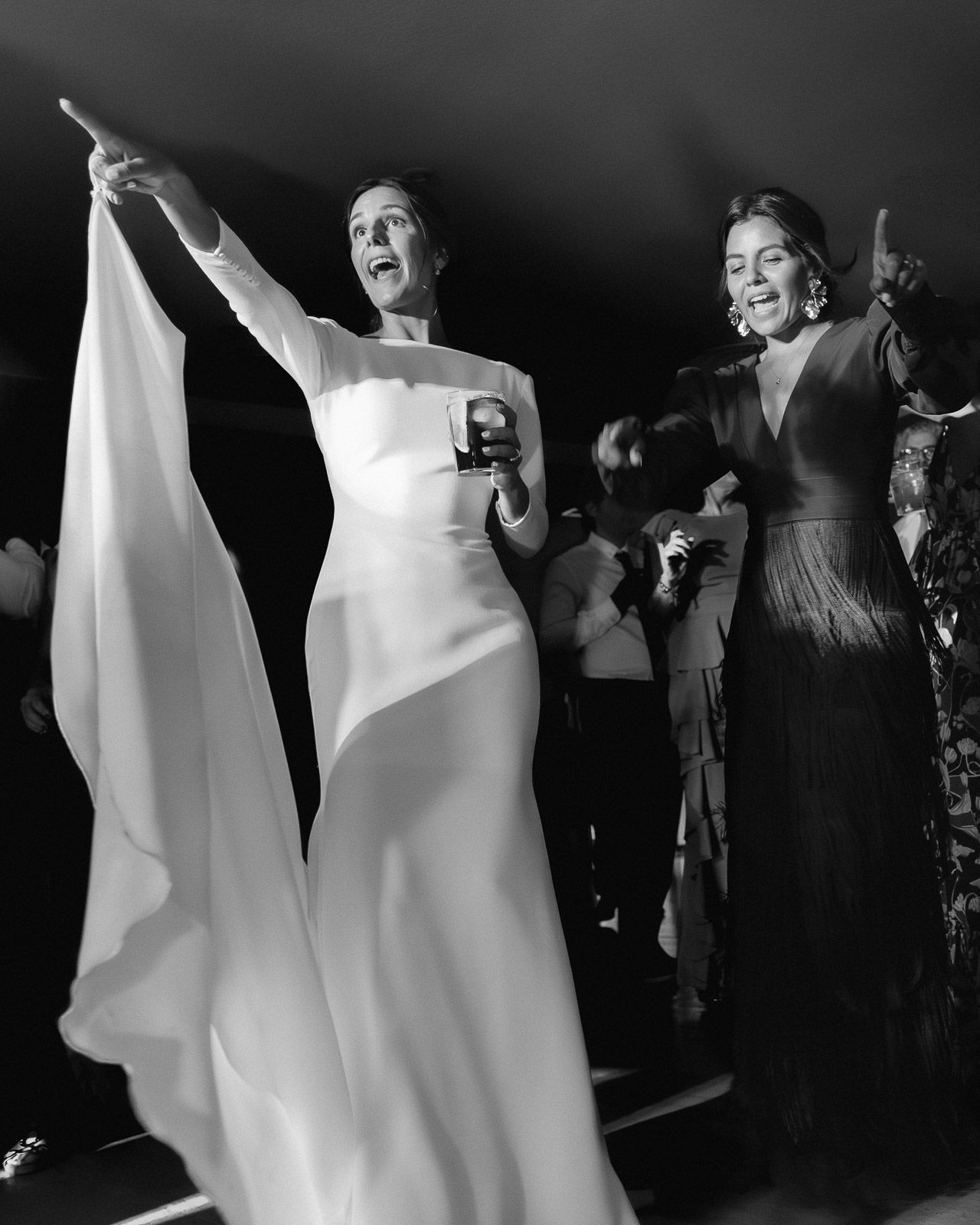 Two elegant women in formal gowns on stage, one in white cape dress pointing, black and white photo.