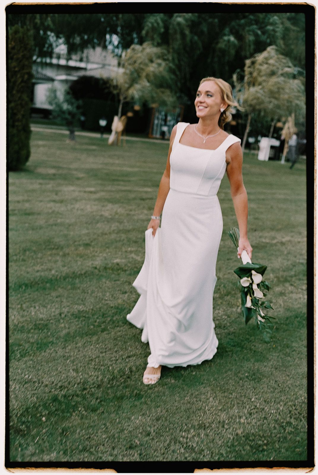 Bride in white gown holding bouquet walks across green lawn, smiling radiantly on her wedding day.
