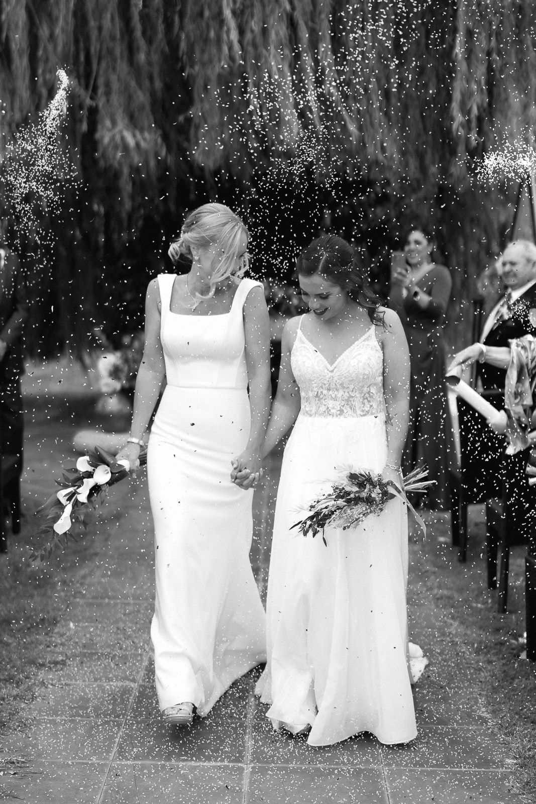 Two brides in white gowns walk hand-in-hand through a confetti shower at their wedding ceremony.