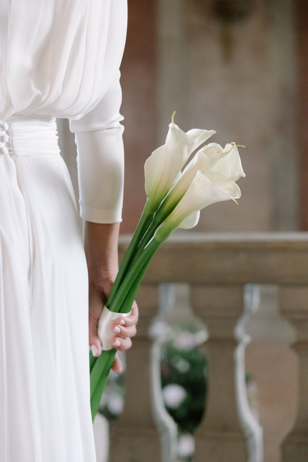 Close-up of bride in white dress holding elegant white calla lily bouquet at outdoor venue.