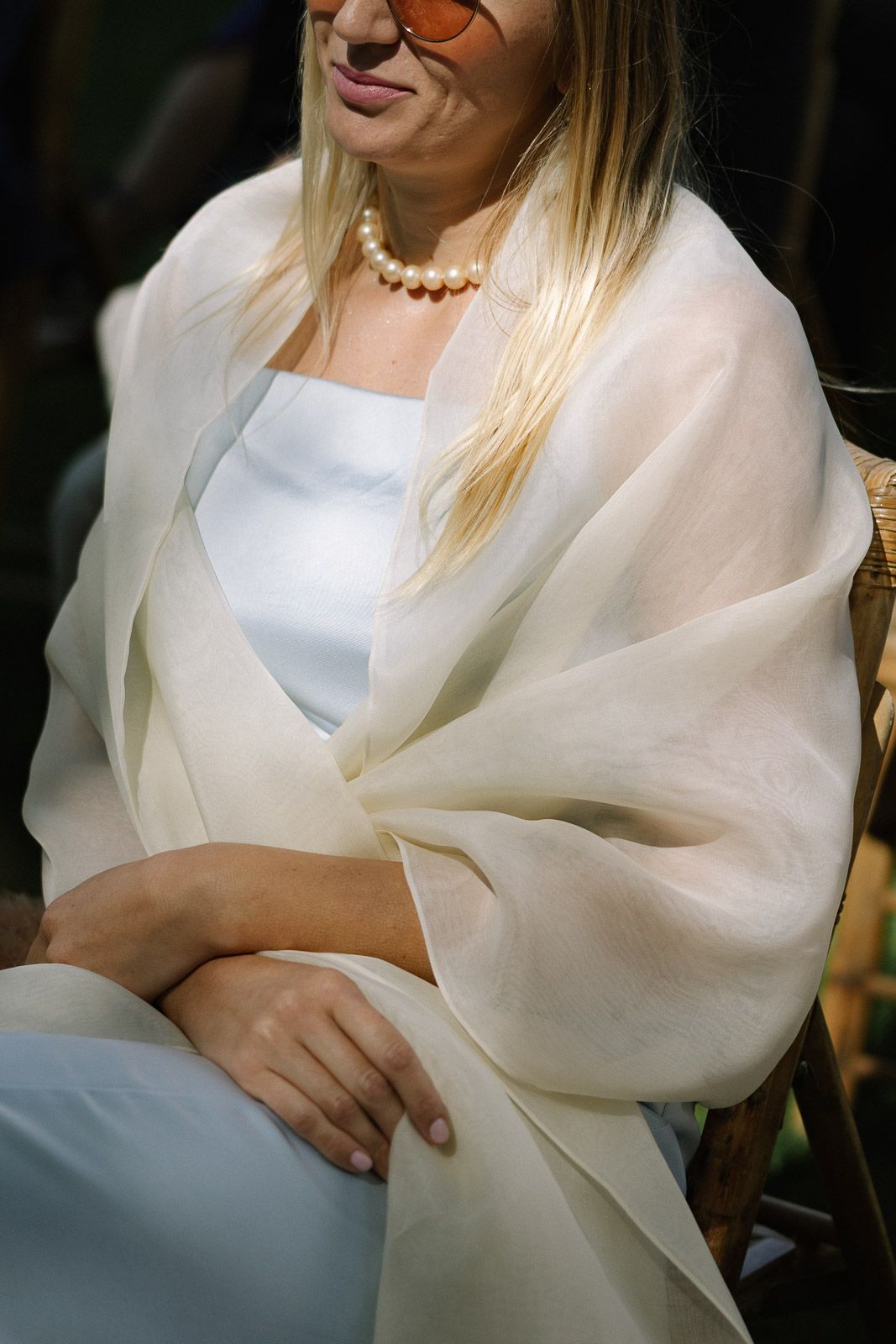 Blonde woman in a light blue dress with a white sheer wrap and pearl necklace seated outdoors.