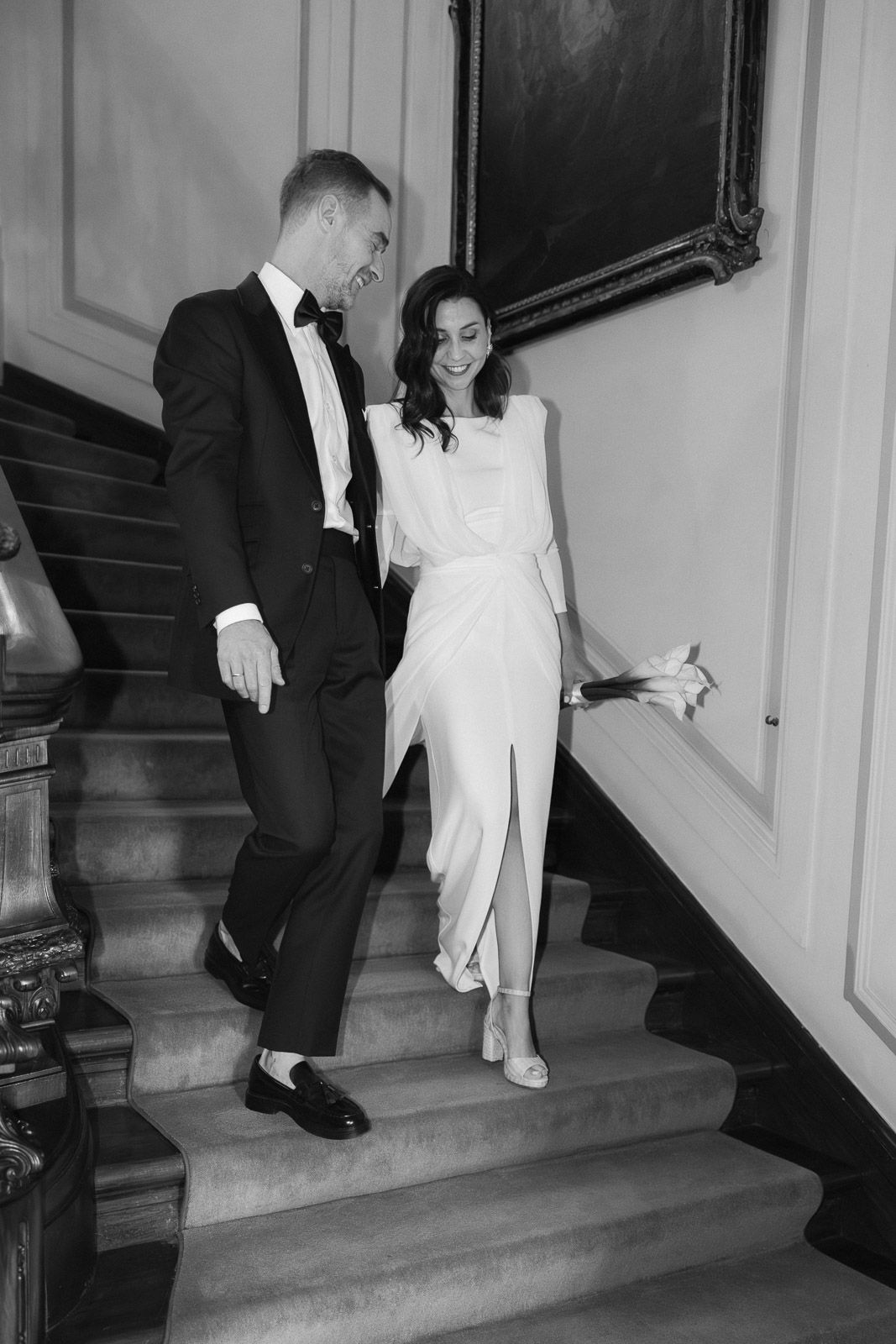 Elegant bride and groom in black and white, descending grand staircase in formal wedding attire.