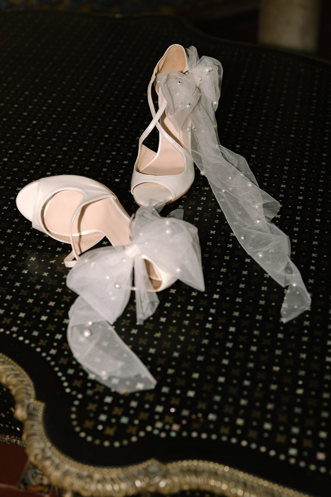 Elegant white bridal heels with sheer tulle ribbon bows displayed on a dark sequined surface.