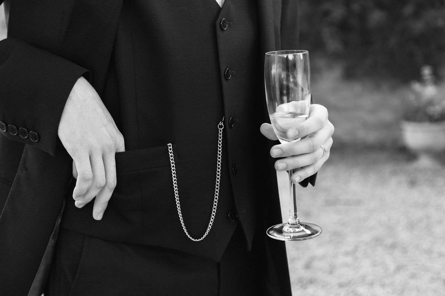 Black and white photo of a groom holding champagne flute, wearing a suit with pocket watch chain at a wedding.