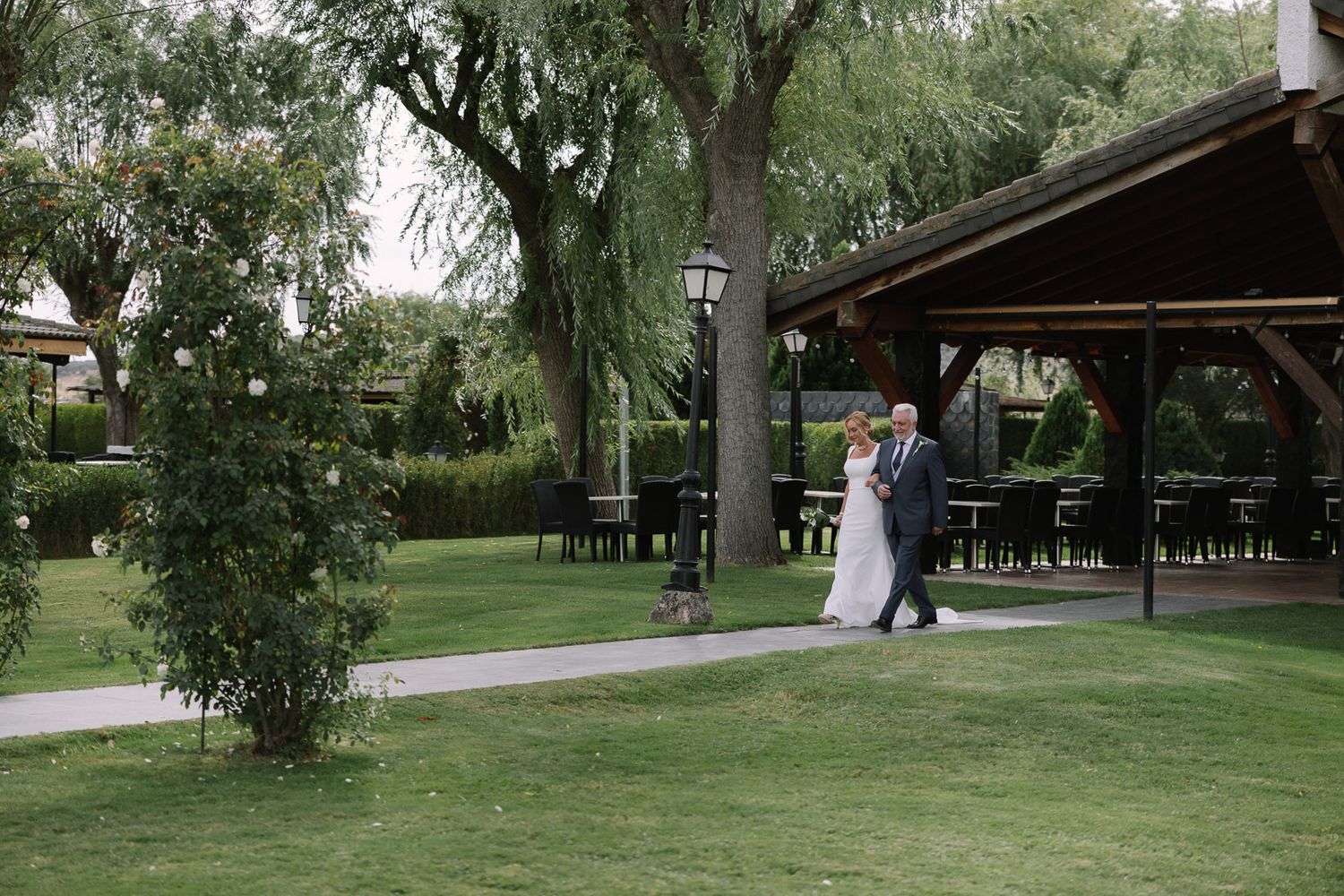 Bride and groom walking hand in hand in a lush green garden near an elegant outdoor pavilion.