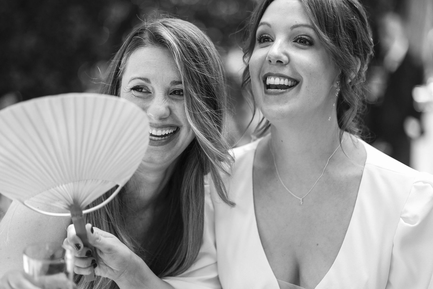 Two joyful women laughing at a wedding, one holding a white paper fan, captured in elegant black and white photography.