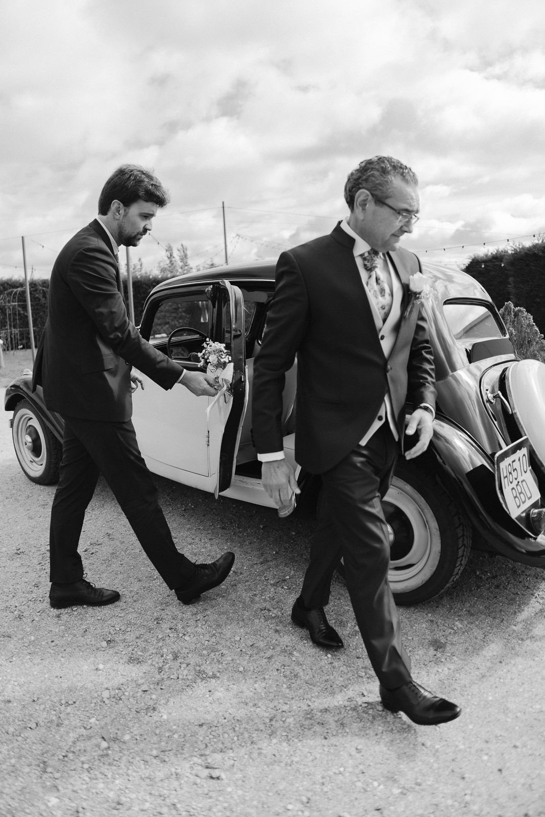 Two men in formal suits exit a vintage black and white car at a wedding ceremony outdoors.