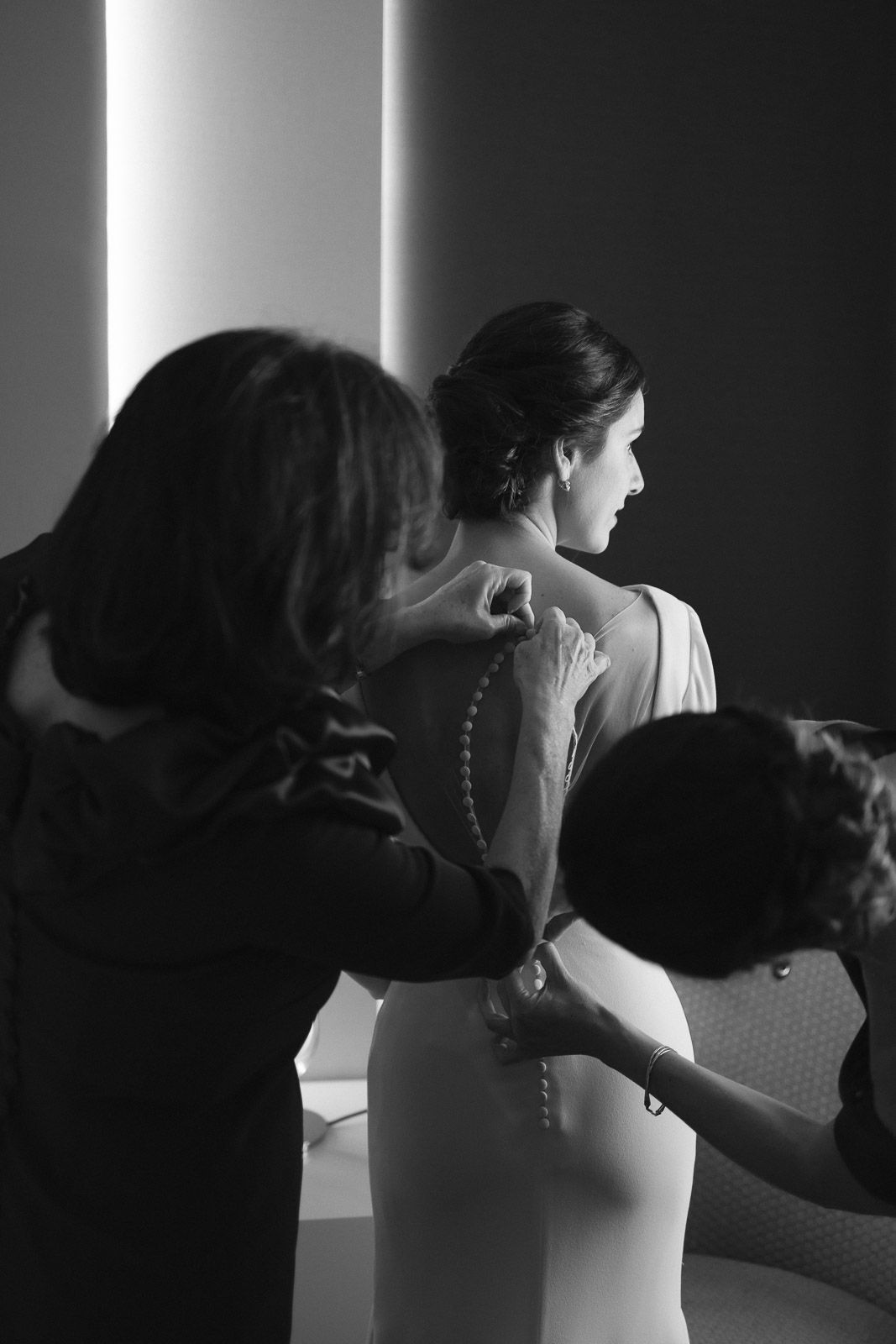 Black and white photo of bridesmaids helping bride with pearl necklace and wedding dress buttons before ceremony.