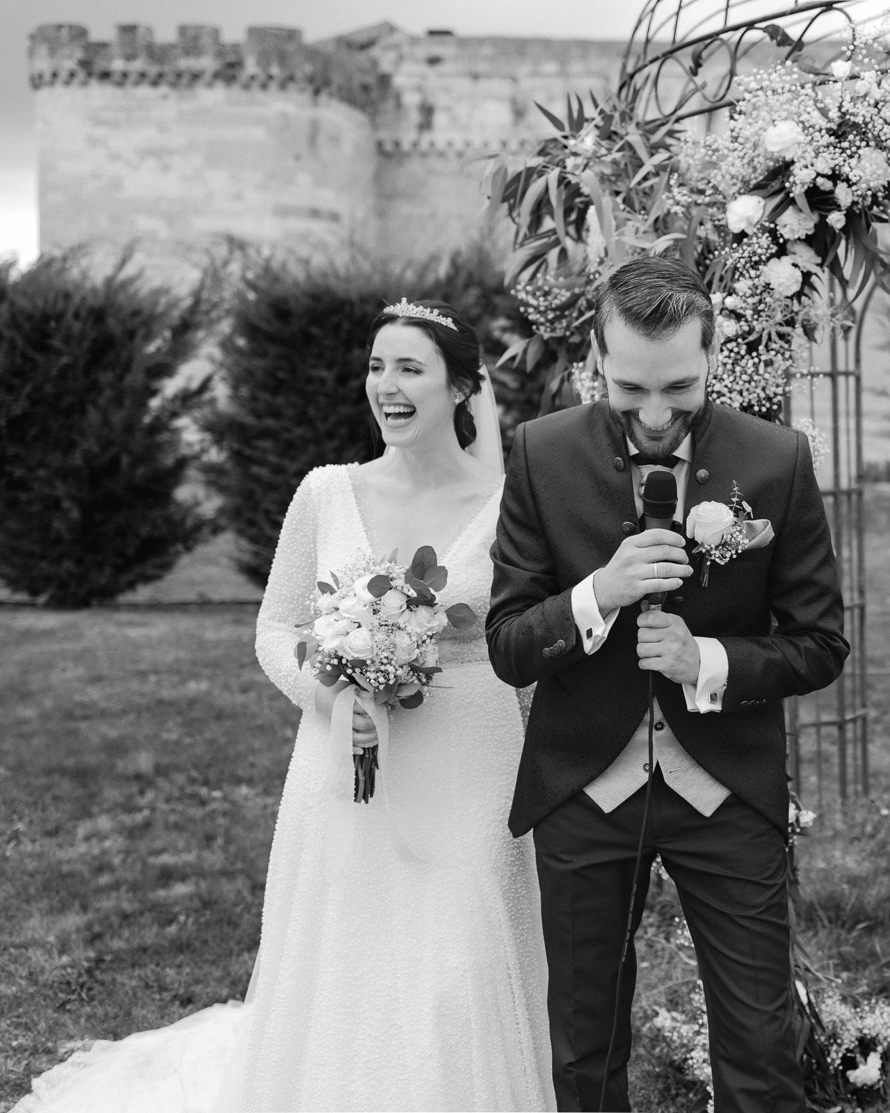 Black and white photo of a laughing bride in a lace gown and groom in a suit at an outdoor castle wedding ceremony.