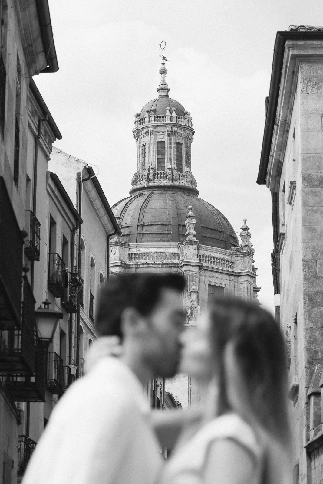 Black and white photo of a couple almost kissing with a historic domed church tower rising between old European buildings.