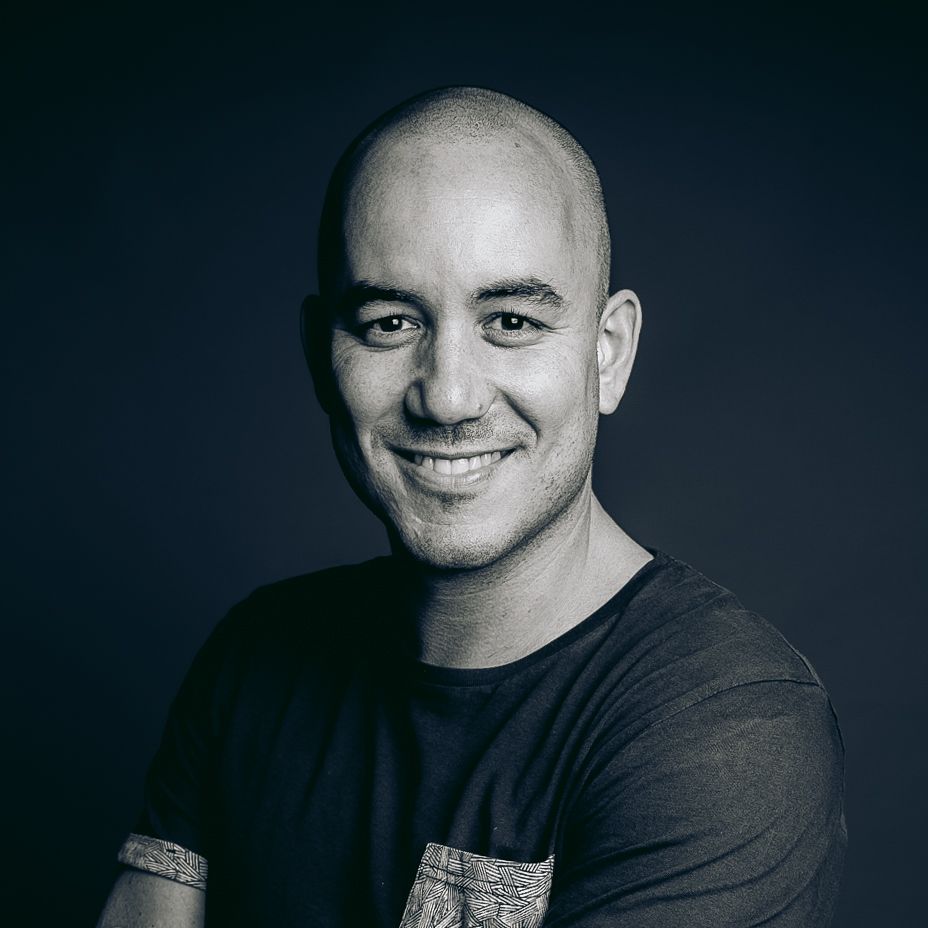 Bald man with a warm smile wearing a dark t-shirt, photographed against a dark studio background.