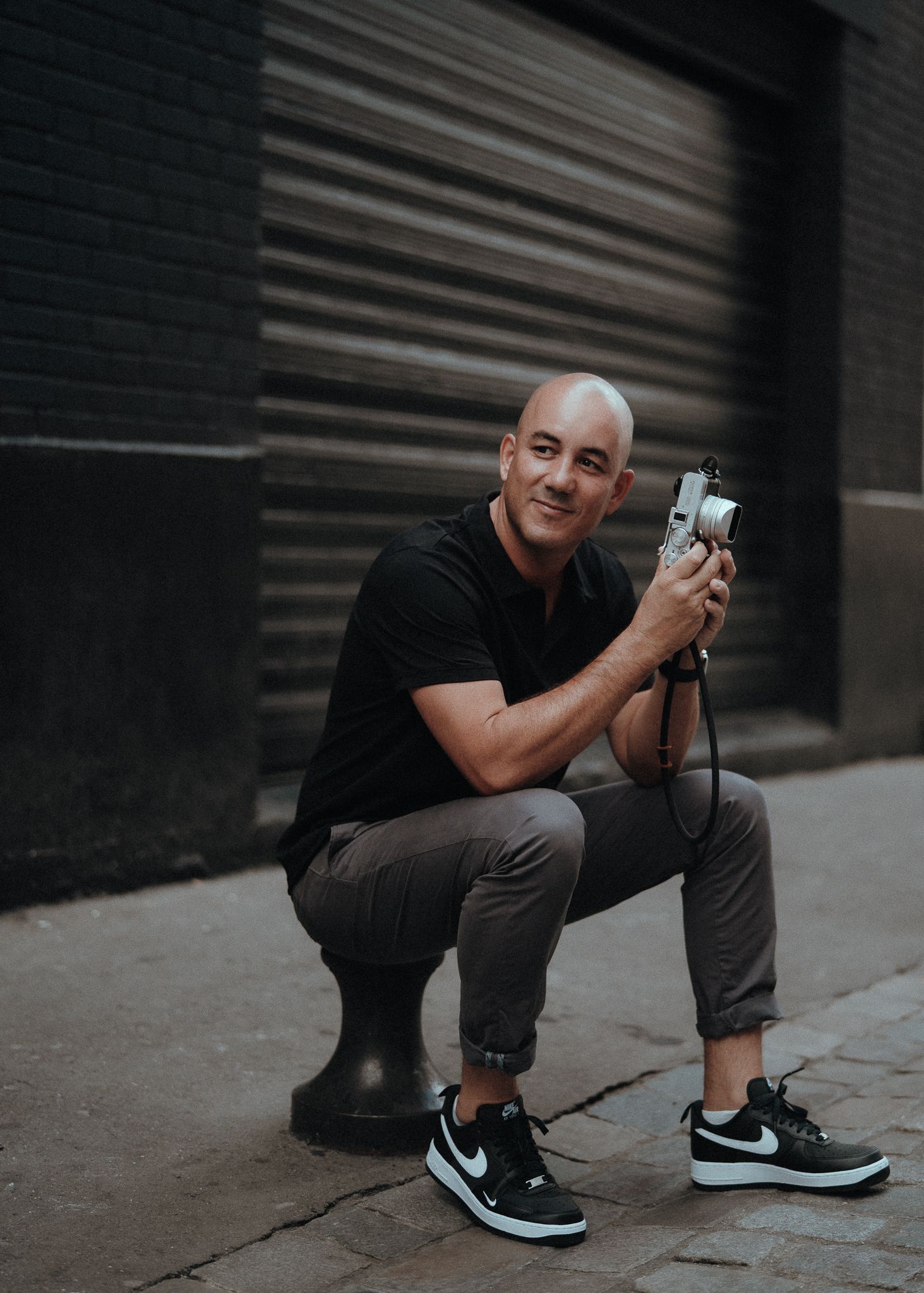 Bald man in black shirt crouching on city sidewalk holding a camera, smiling, wearing Nike sneakers.