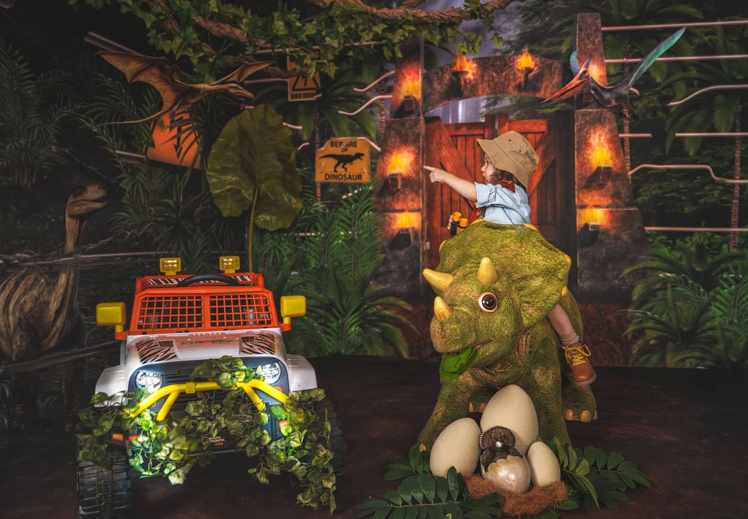 Two-year-old boy in Jurassic Park outfit sitting on a toy T-rex pointing to another dinosaur in L Buchanan Photography photo session Perry H