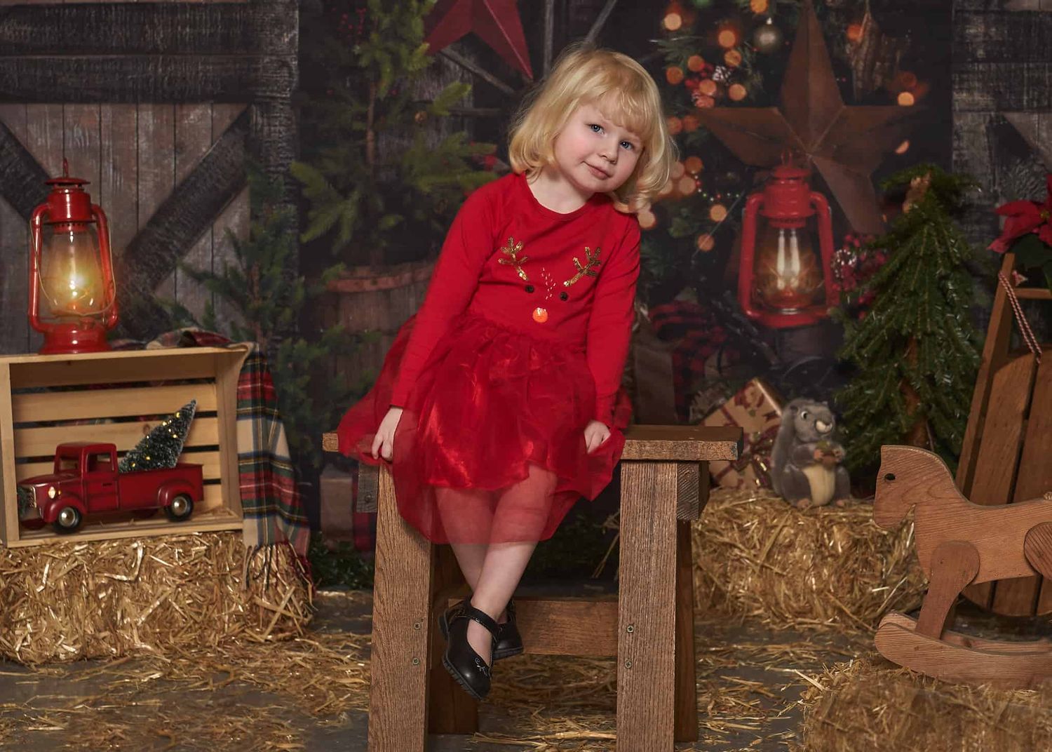 Five-year-old blonde-haired girl in red Christmas dress sitting on wooden bench in barn setting