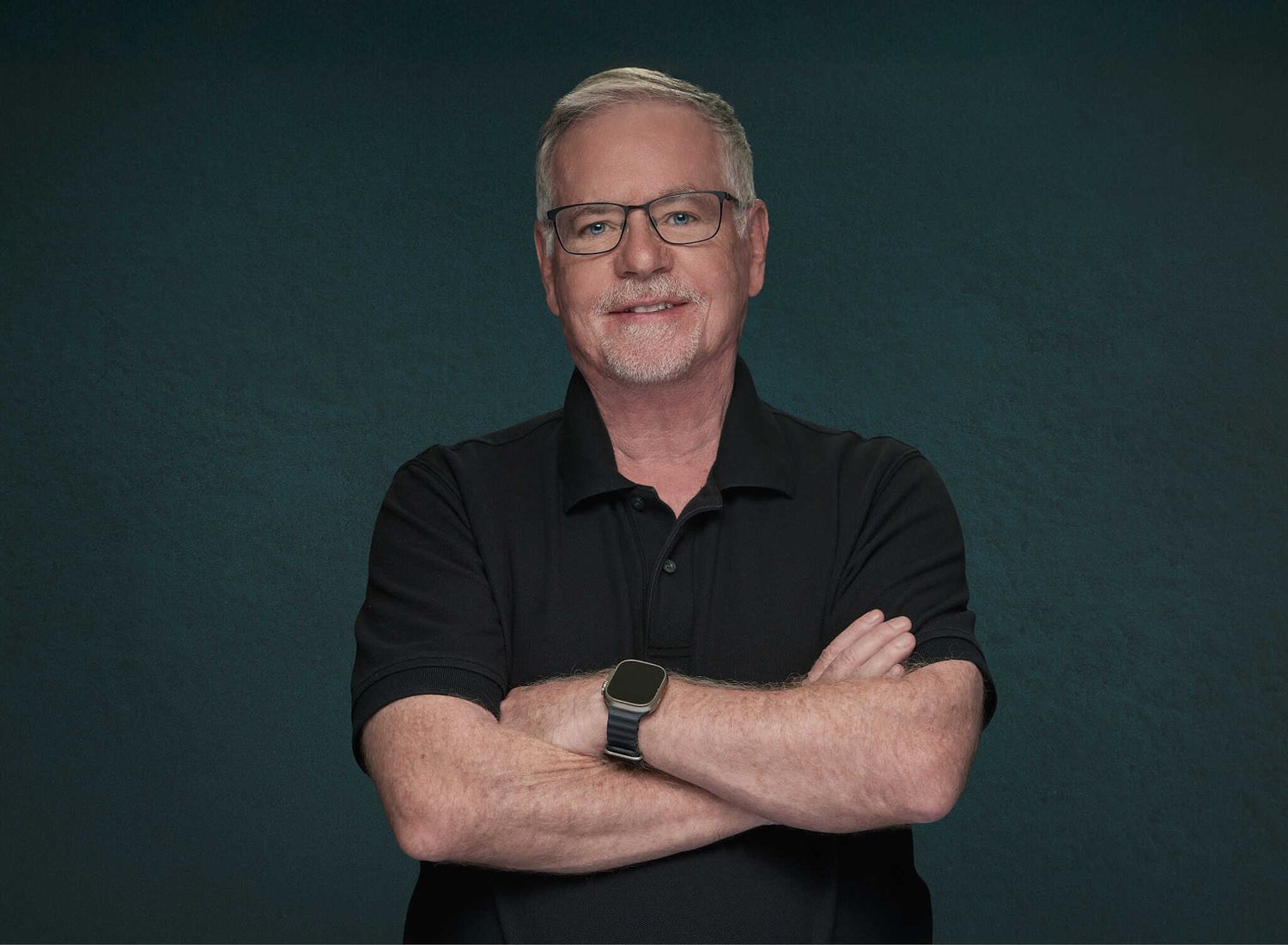 Gray-hair ikder gentleman in black polo shirt and black-rimmed glasses with arms crossed showing a relaxed, happy smile.