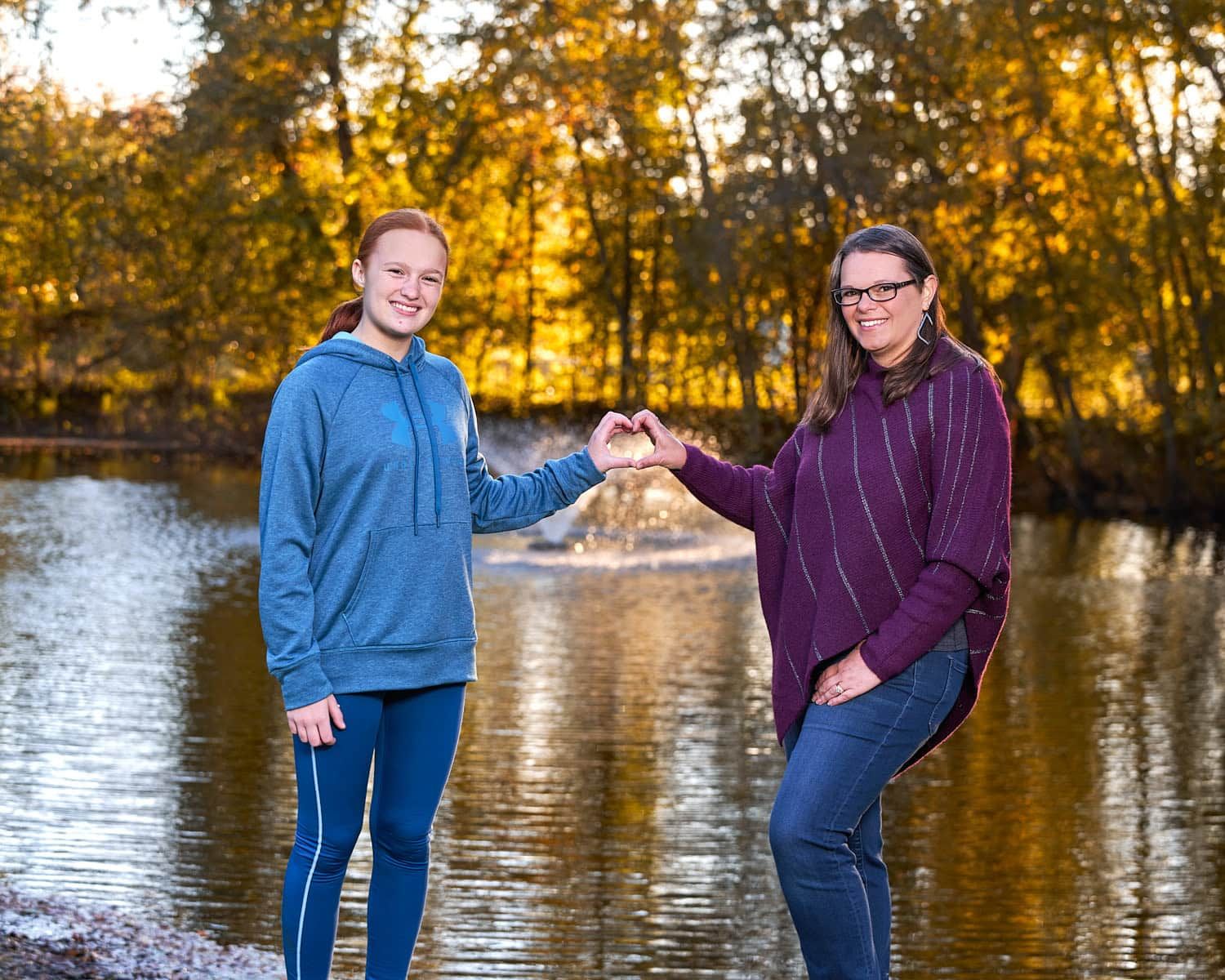 Mother and tween daughter making a heart symbol with their combined hands standing in front of a pond.