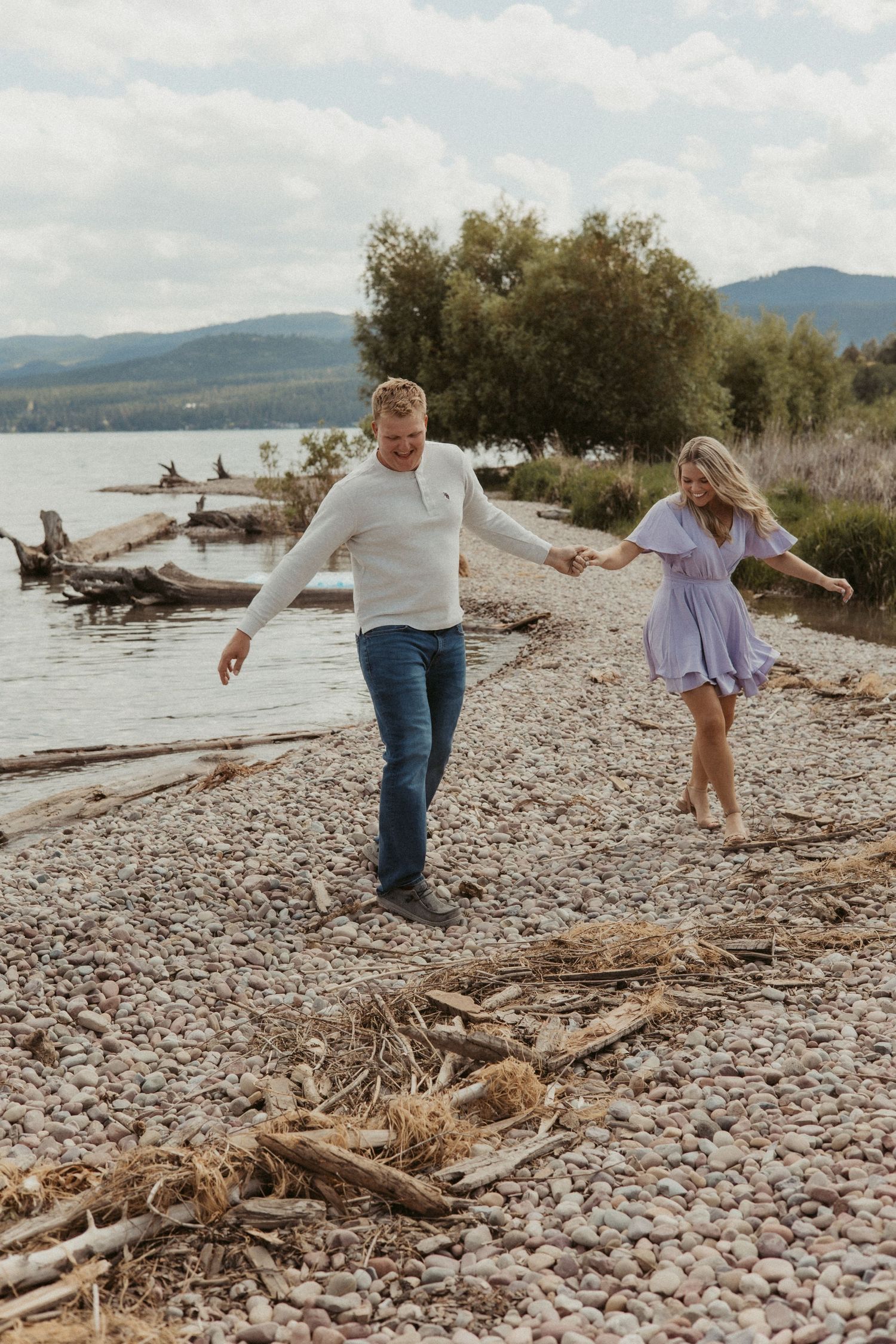 Couple walking playfully along a rocky beach shore with mountains and driftwood in the background.