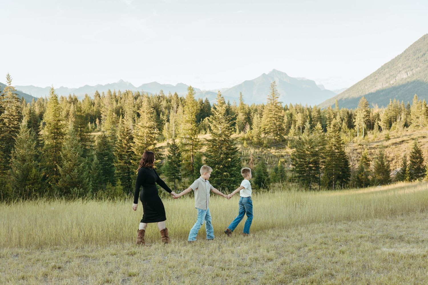 Three people holding hands and dancing in a meadow with evergreen trees and mountains in the background.