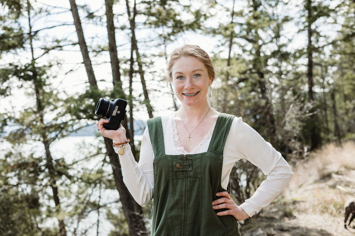 A woman in a green dress holds a camera while standing in a forested area.