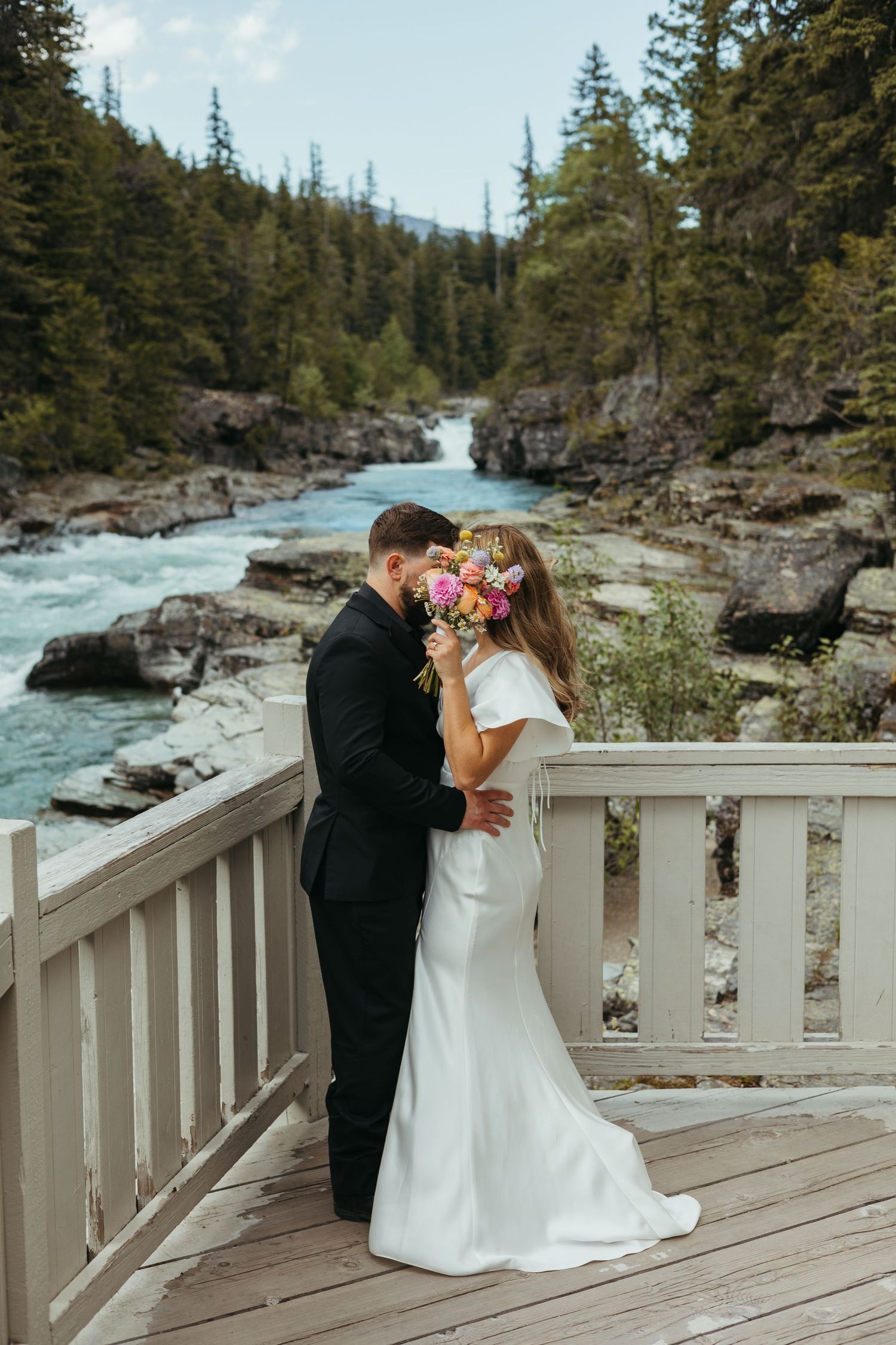 Bride and groom embrace on wooden bridge with colorful bouquet, framed by rushing river and pine forest backdrop.
