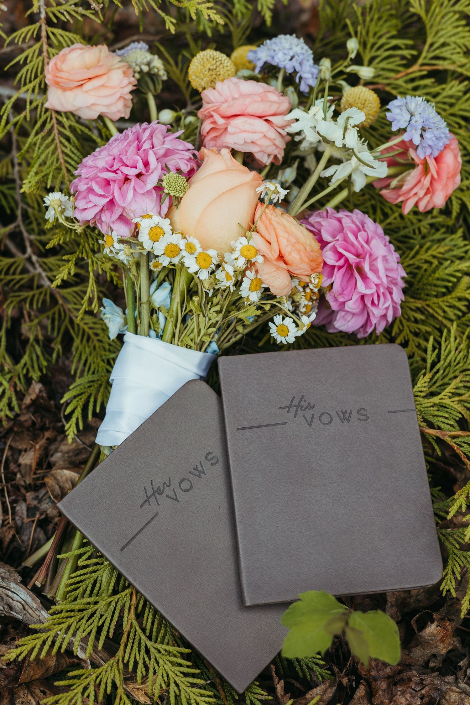 Two gray vow books labeled His and Her Vows rest beside a colorful bridal bouquet with pink dahlias and daisies.