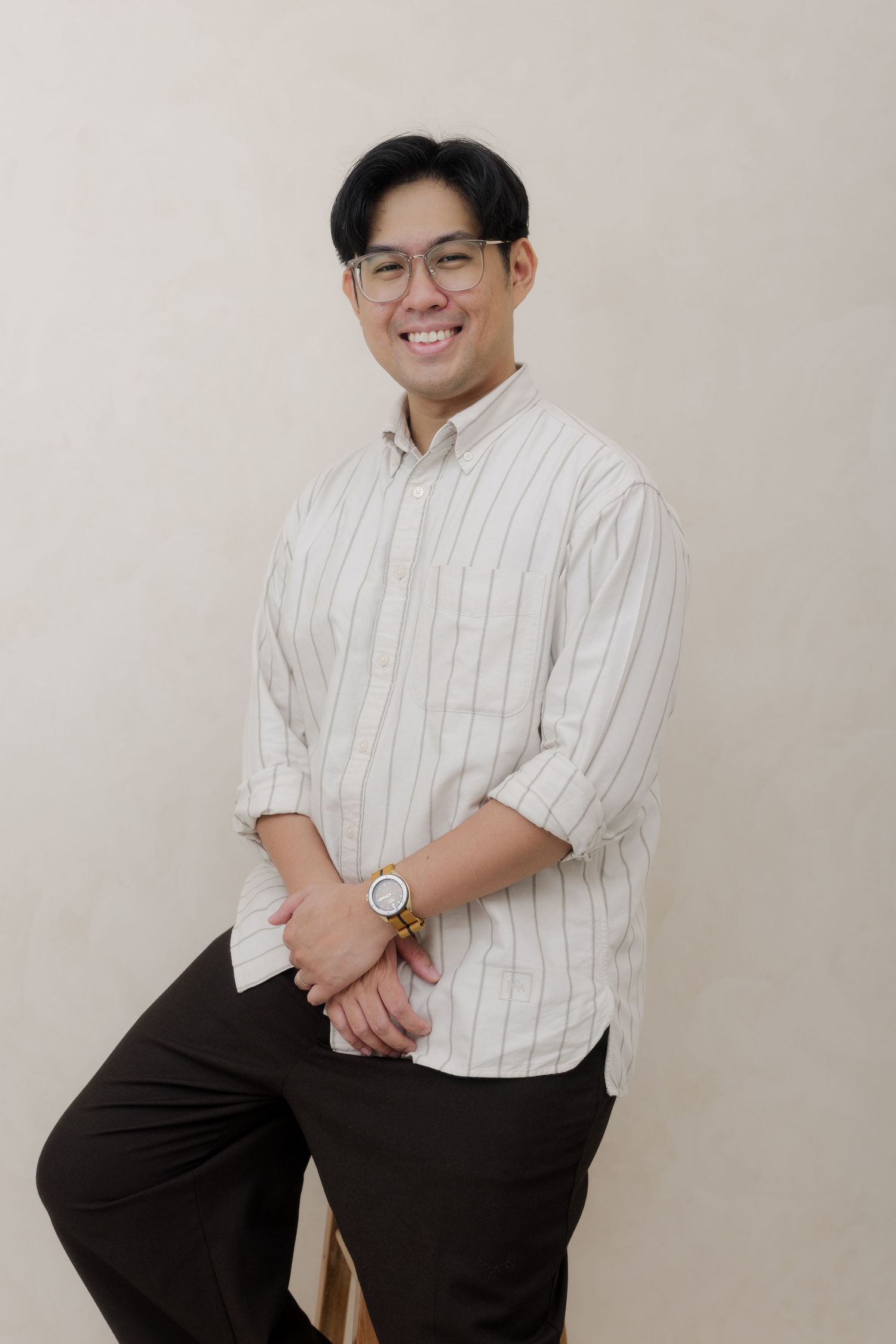 Smiling young Asian man in striped shirt and glasses posing casually against a neutral background.