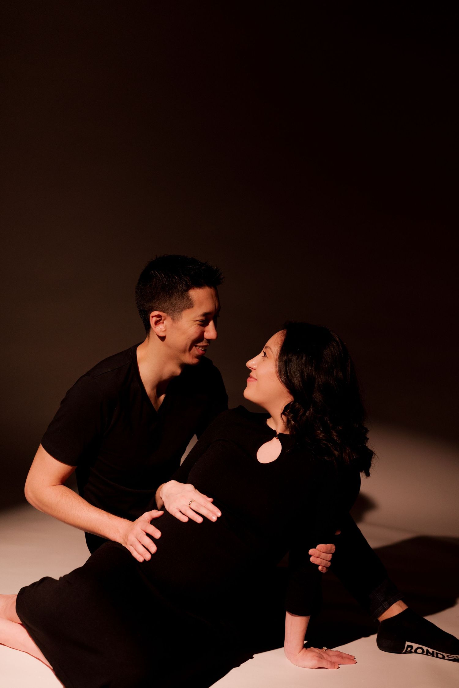 A couple in black outfits sit together on the floor, smiling at each other, with the woman visibly pregnant.