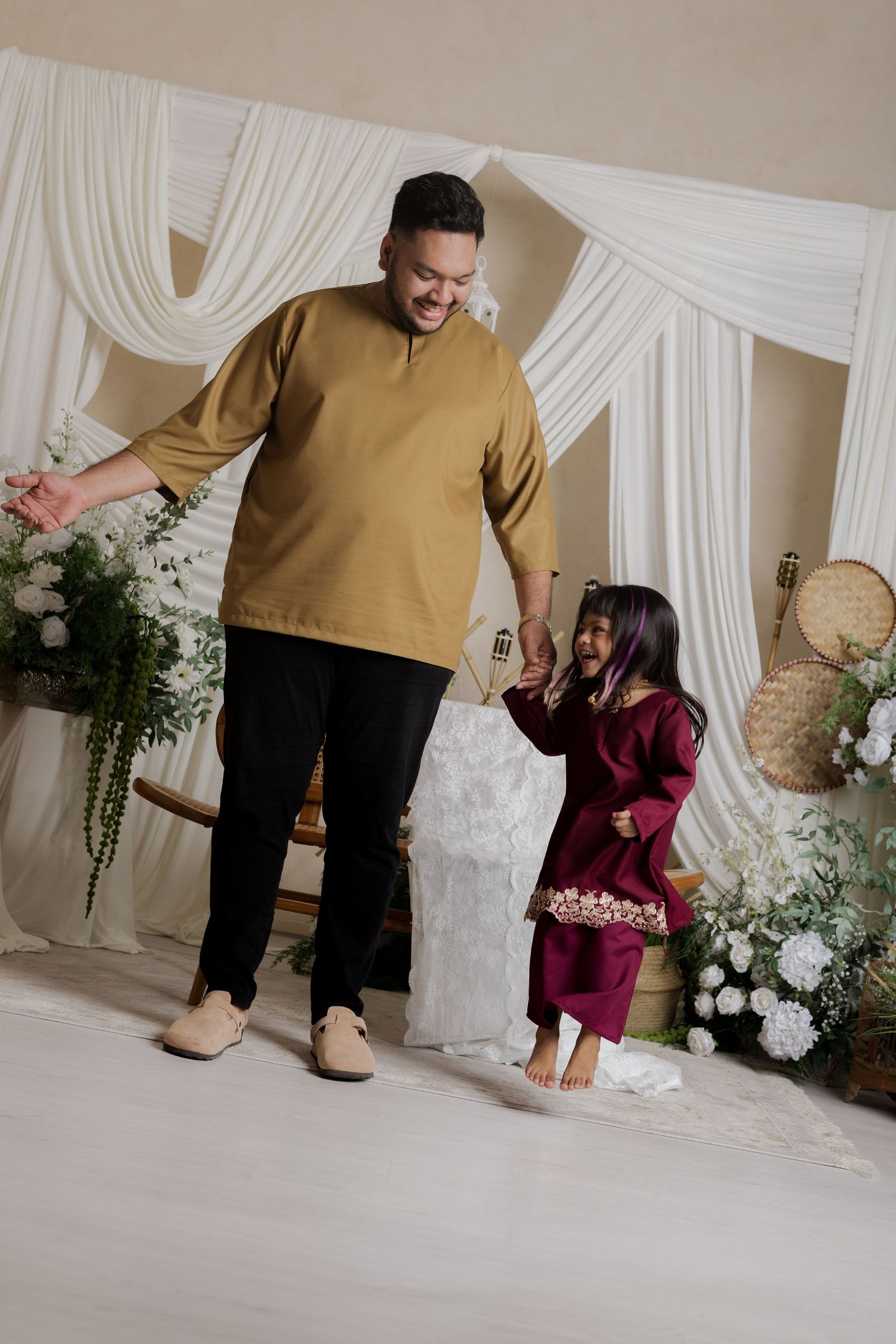 Man in gold baju kurung dancing with young girl in maroon outfit at decorated Eid celebration backdrop.